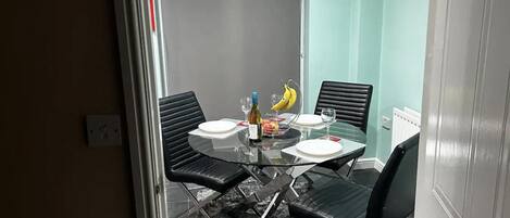 Un comedor con mesa de vidrio, cuatro sillas negras, una botella, un vaso y un bol de fruta.