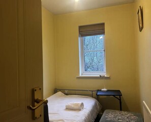 Una habitación pequeña con cama individual, ventana, mesita de noche y puerta.