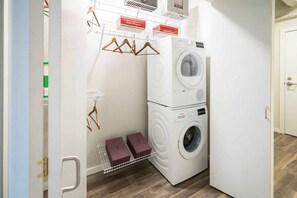 A compact laundry room with a stacked washer and dryer, a drying rack, and a small shelf with boxes. - Chic Downtown Living—Sleek Apt Near Attractions (Cleveland)