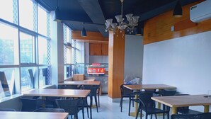 A cafe with wooden tables and black chairs, a coffee machine, and large windows. - Hotel City View (Dhaka)