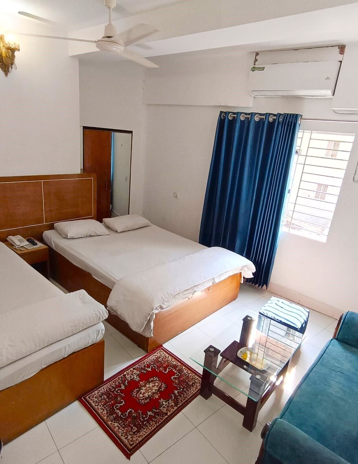 Habitación de hotel con dos camas, un ventilador de techo, un equipo de aire acondicionado, una cortina azul, una mesita de centro de vidrio y un tapete rojo estampado.