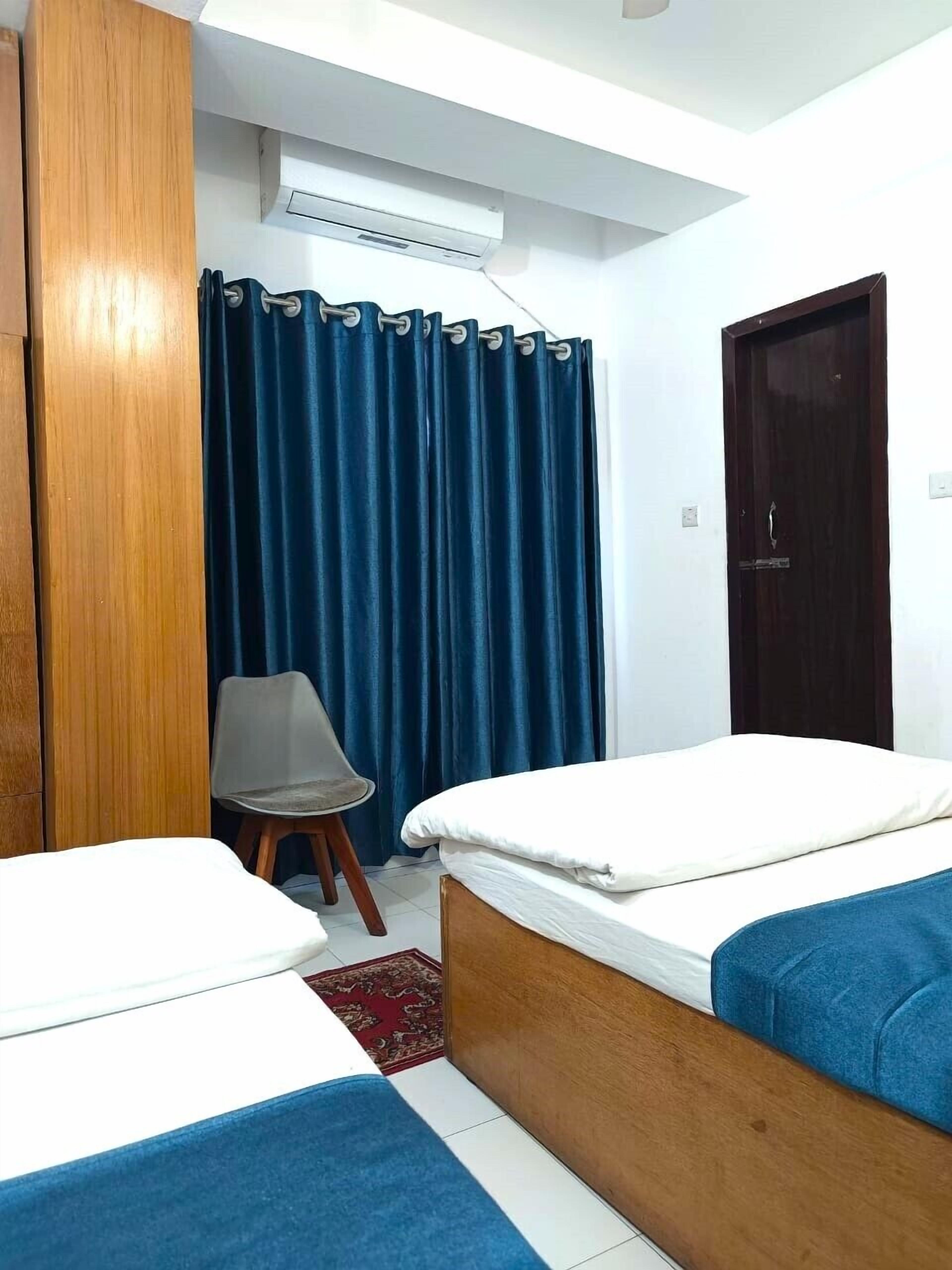 Habitación de hotel con dos camas individuales, un armario de madera, una silla, una cortina azul y un equipo de aire acondicionado.