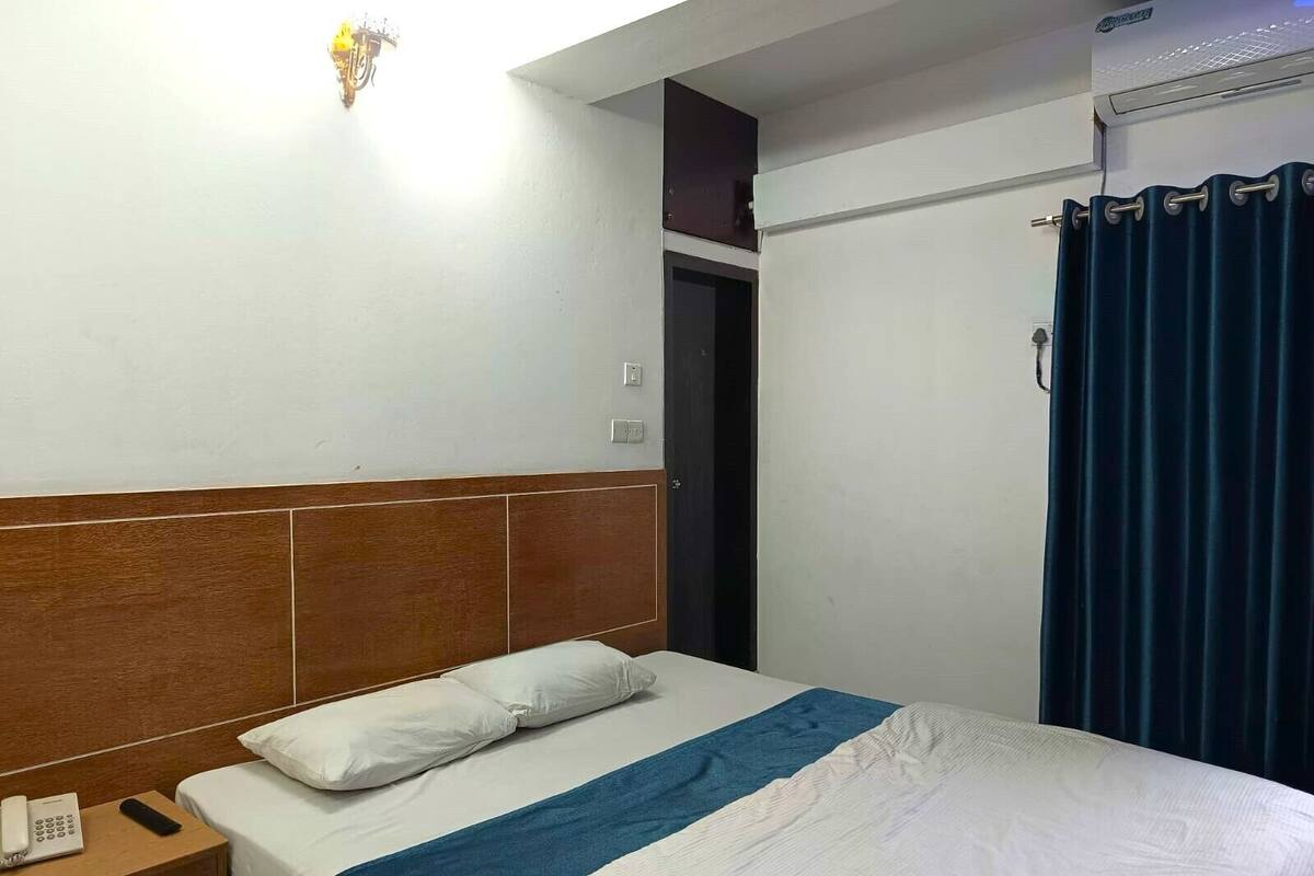 A hotel room with a wooden headboard, a bed with white and blue bedding, a bedside phone, and a curtain.