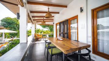 Enjoy meals in this spacious dining area with a long table, comfortable chairs, and a large window offering a view of the outdoor patio and lush greenery.