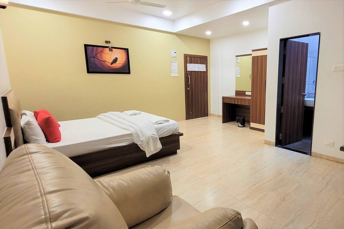 A hotel room with a bed, a sofa, a ceiling fan, a painting, a bathroom with a mirror, and a door leading to another room.
