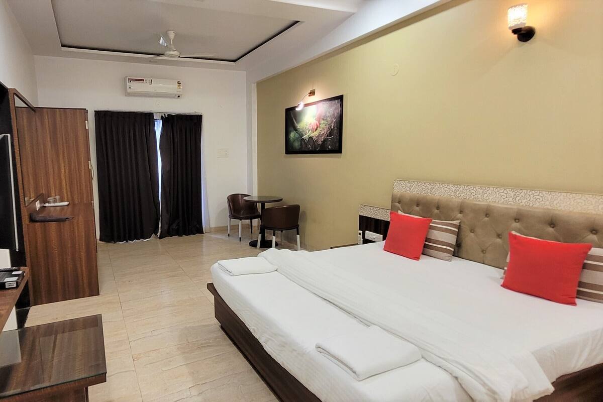 A hotel room with a large bed, a desk, a chair, a small table, a wall-mounted air conditioner, and a door.