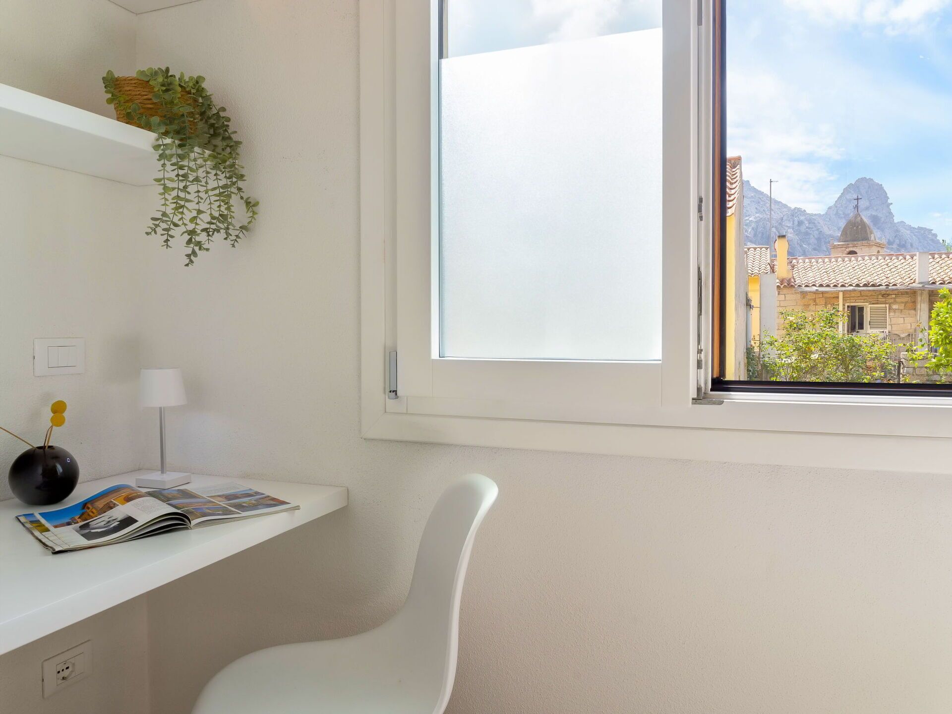 A modern, minimalist room with a white desk, a white chair, a window with a view of a mountain and traditional architecture.