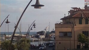 Marina costeira com barcos atracados, estrada que leva a um edifício com placa vermelha e postes de luz.
