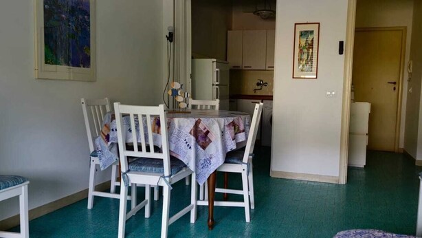 Uma sala de jantar com uma mesa posta para quatro pessoas, rodeada por cadeiras brancas com almofadas azuis. Uma cozinha é visível ao fundo, e uma porta dá para outro cômodo.