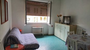Um quarto pequeno e acolhedor com sofá, mesa e cadeiras, armário e janela com persianas.