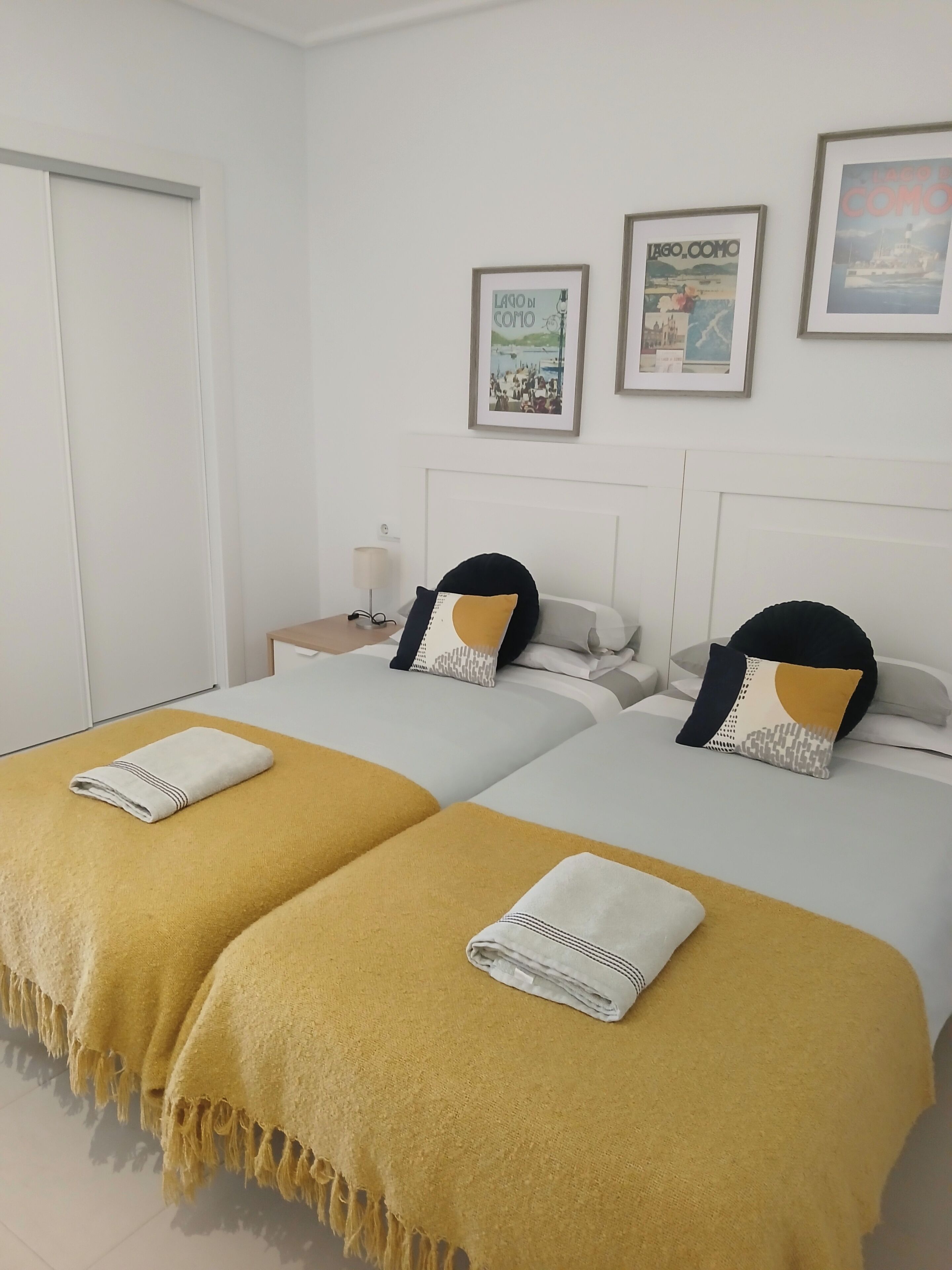 A hotel room with two beds, each with a yellow blanket and a white towel. There are framed pictures on the wall, a nightstand with a lamp, and a door to the left.