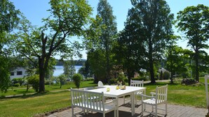 Eine Terrasse im Freien mit einem weißen Tisch und Stühlen, umgeben von üppiger Bepflanzung und Blick auf einen See.