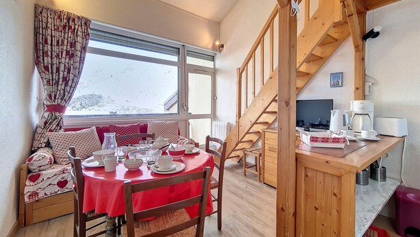 A cozy dining area with a red tablecloth, wooden chairs, and a view of a snowy landscape through large windows. - Cozy apartment near slopes with parking (Les Menuires)