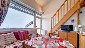 A cozy room with a dining table set for two, a sofa with patterned cushions, a television, and a kitchen area with a coffee maker. - Cozy apartment near slopes with parking (Les Menuires)