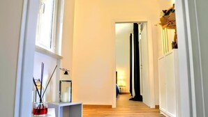 Un corridoio stretto con pavimento in legno, pareti bianche e uno scaffale con una candela e un diffusore di fragranze.