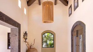 Un pasillo con techo alto, vigas de madera, un candelabro colgante, un jarrón grande con ramas, una puerta con paneles de vidrio y una ventana con vistas a la vegetación.