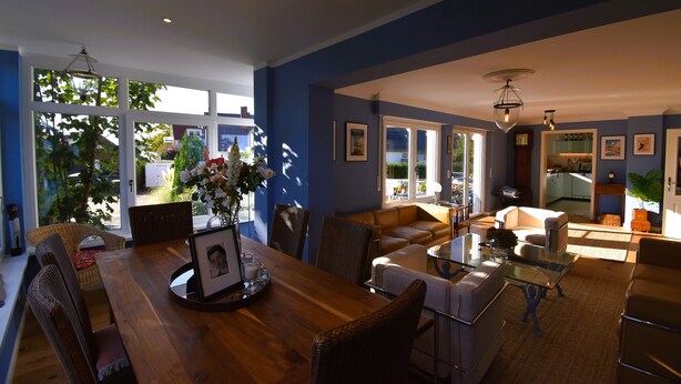 A spacious living room with a wooden dining table, comfortable seating, and large windows offering a view of the outdoors. - Sunny 4-room garden apartment with pool near the lake for up to 6 people (Eriskirch)