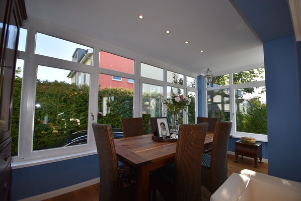 A dining area with a wooden table, wicker chairs, and a view of greenery and buildings through large windows.