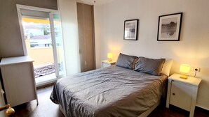 Une chambre à coucher avec un lit, deux tables de chevet avec des lampes, une fenêtre donnant sur des immeubles et deux tableaux encadrés accrochés au mur.