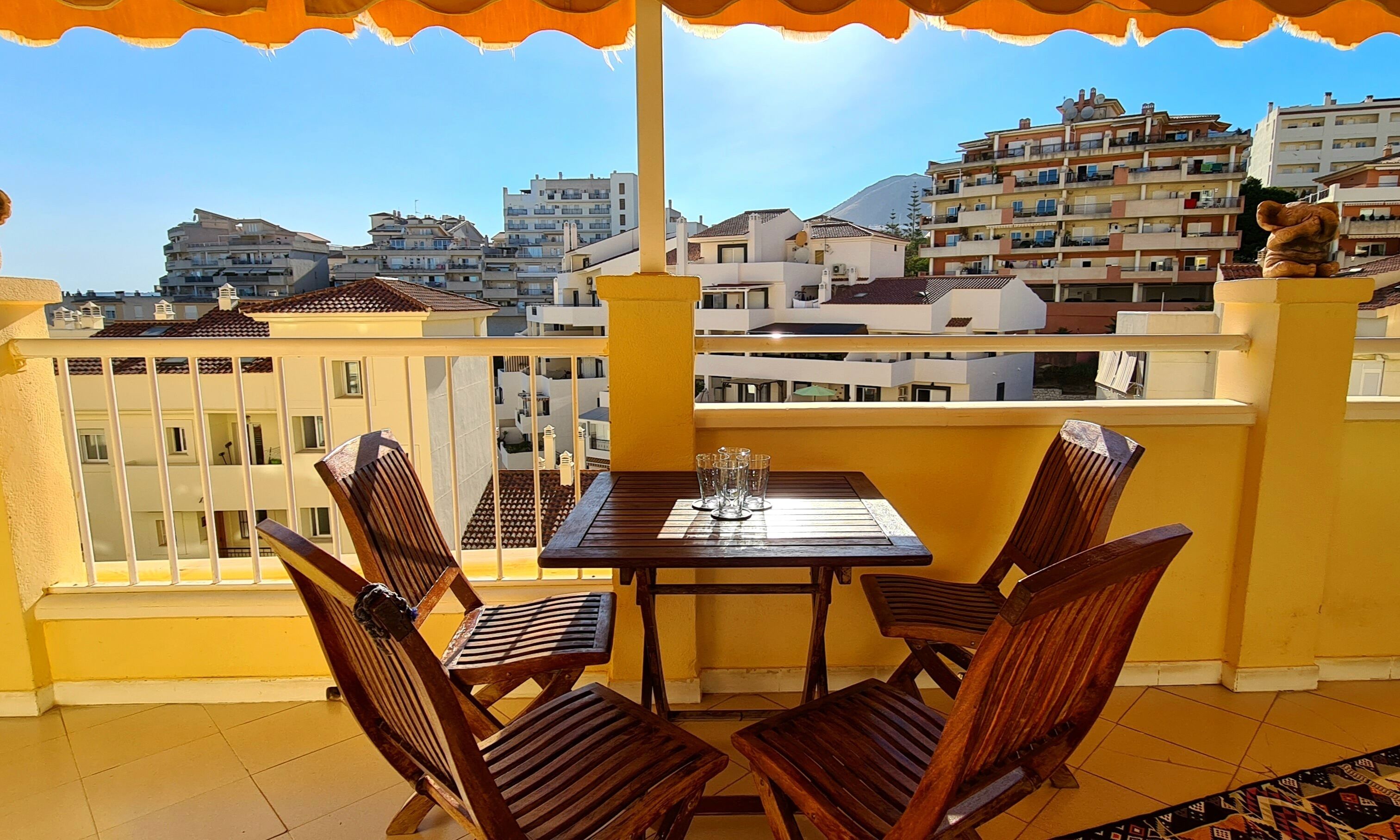 A balcony with a wooden table and chairs, overlooking a cityscape with buildings and a mountain in the distance.