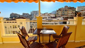 Un balcon avec une table et des chaises en bois, donnant sur un paysage urbain avec des immeubles et une montagne à l’horizon.