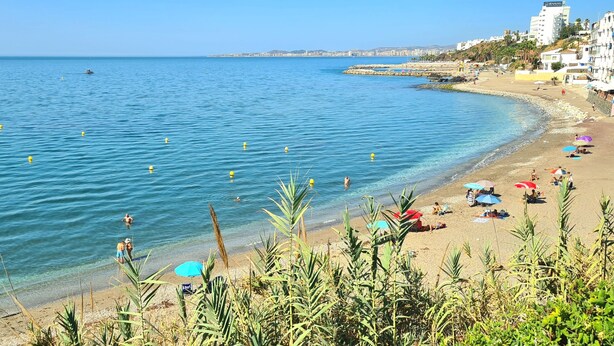Une plage avec une eau d’un bleu limpide, des parasols et des gens.