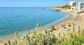 Ein Küstenstrand mit klarem, blauem Wasser, Sonnenschirmen und Menschen.