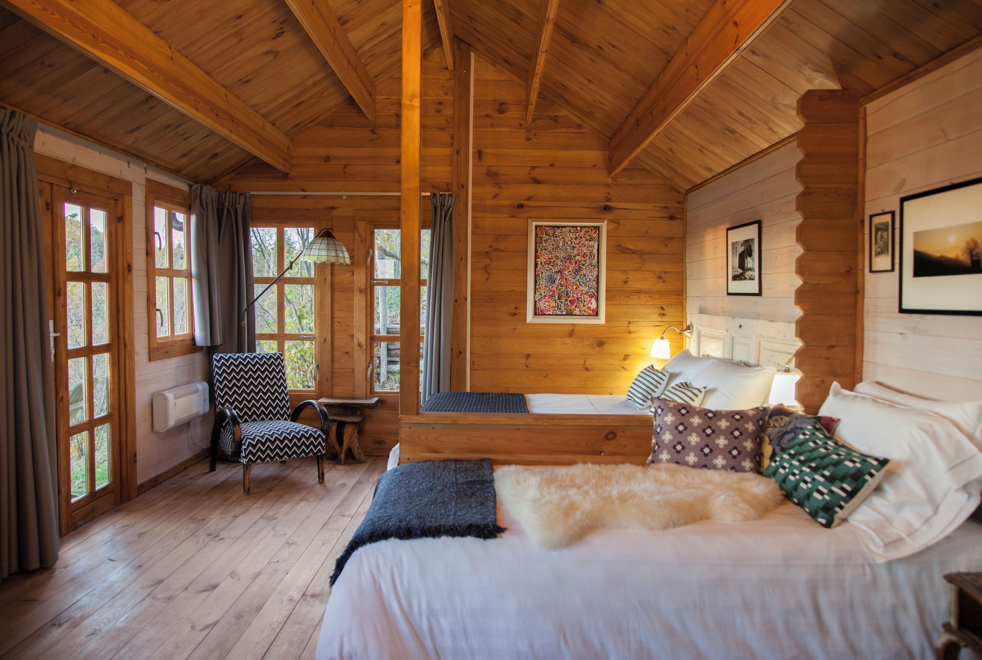 A wooden cabin bedroom with a bed, a chair, a table, and a window with curtains.