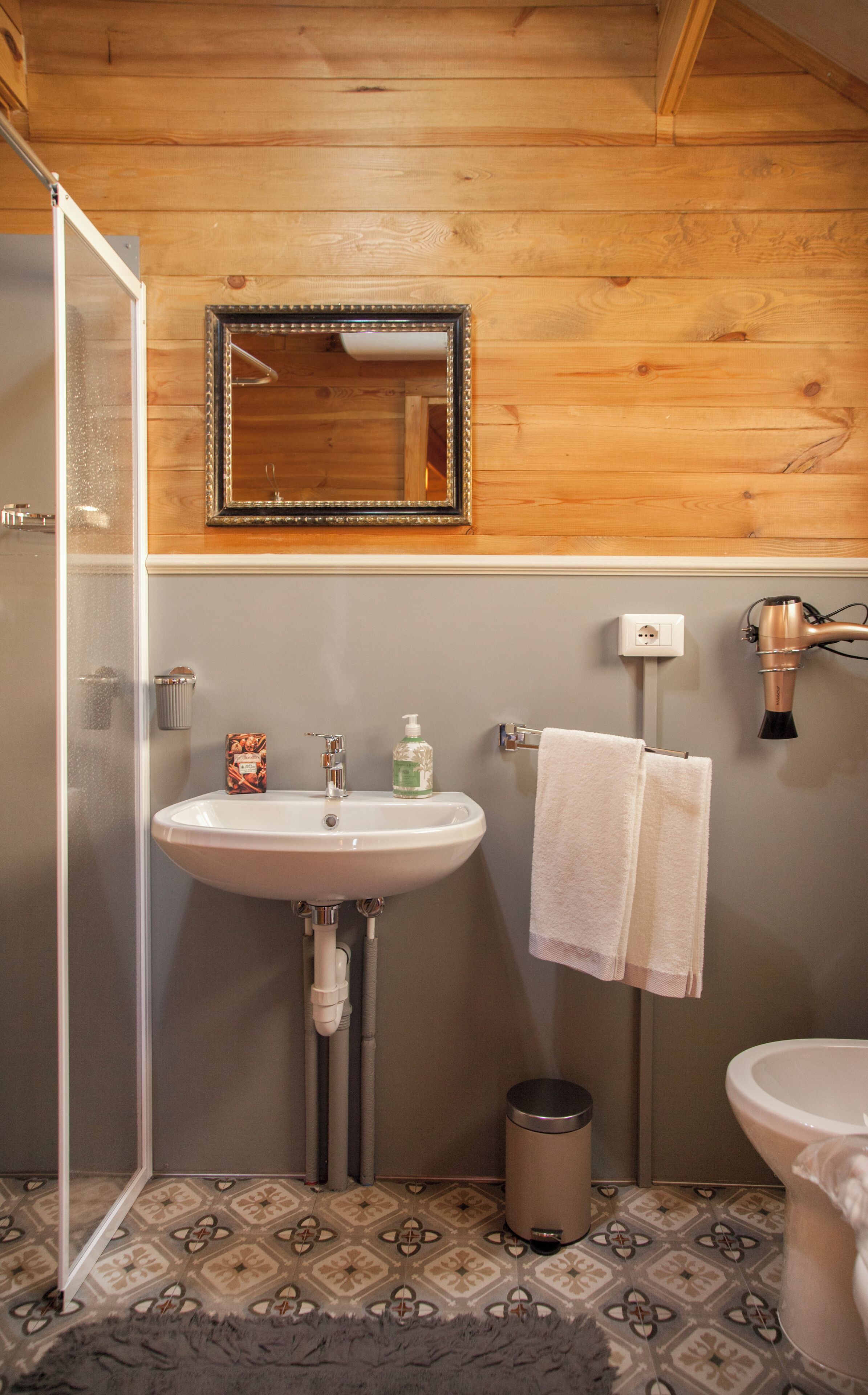Un baño con techo de madera, un lavamanos, un espejo, un perchero para toallas, un secador de pelo y un cesto de basura.