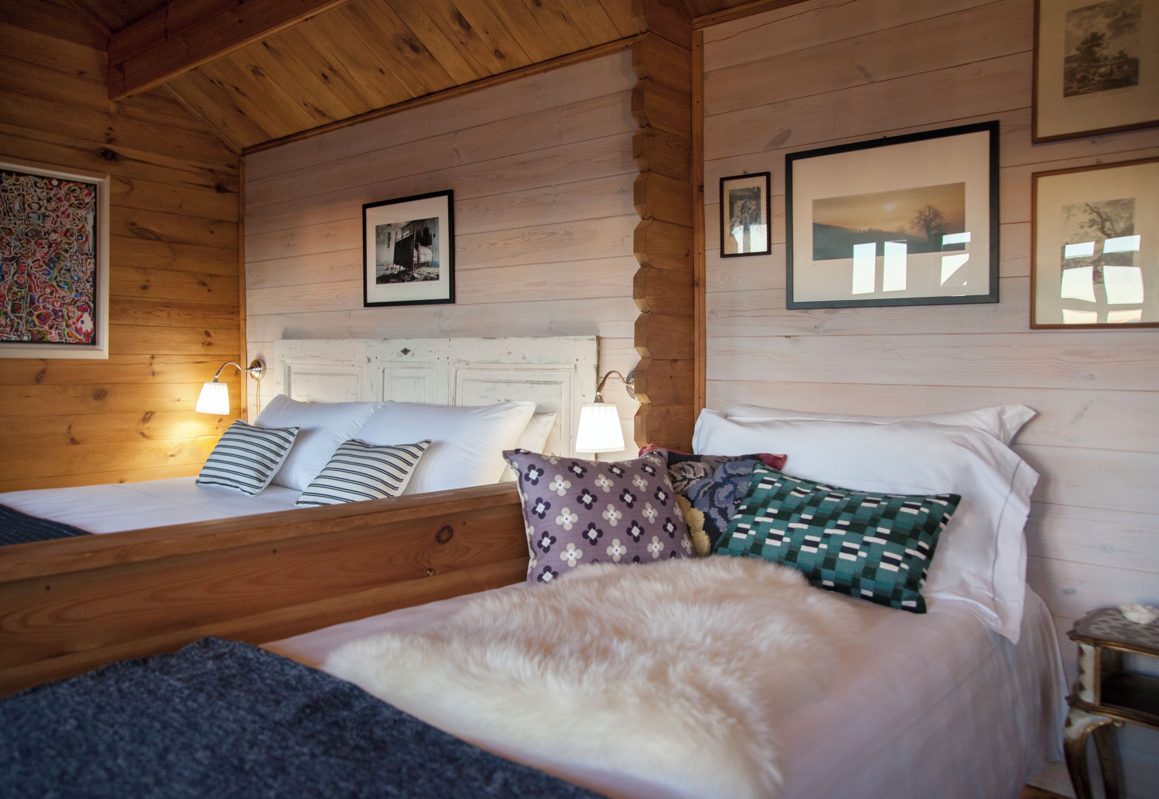A wooden cabin room with two beds, each with different bedding and pillows. There are framed pictures on the walls and a lamp on the left side.