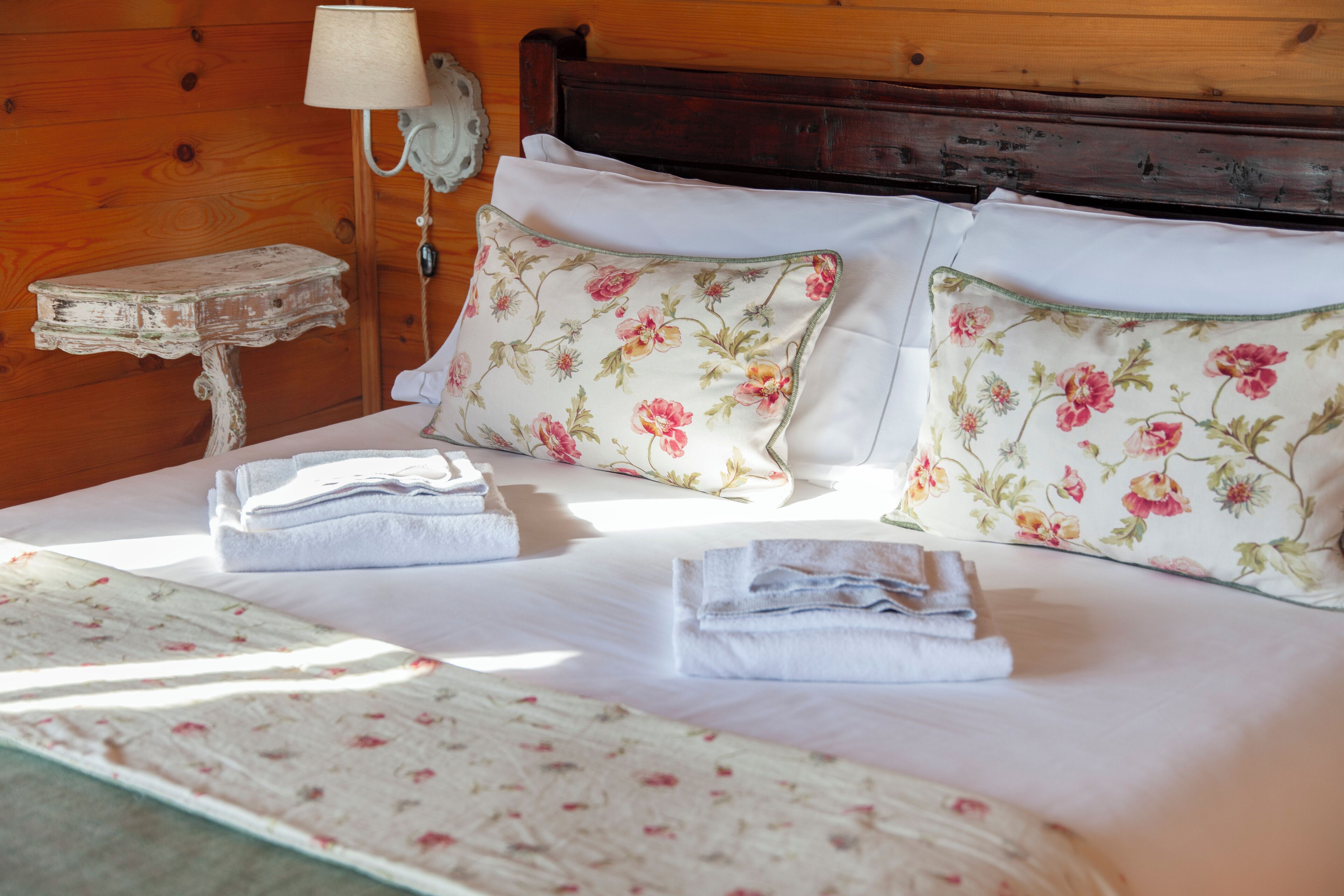 Un lit bien fait, agrémenté d’oreillers à motifs floraux, une petite table de chevet avec une lampe et un téléphone fixé au mur.