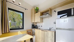 A compact kitchen with a built-in stove, a sink, and a refrigerator. There is a window with curtains, a plant on the counter, and a chair. - Wooden Tiny House with terrace, pool, wifi, pets, parking - 200m from the beach (Jullouville)