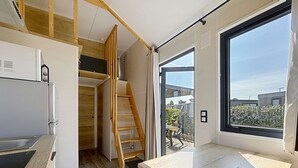 A compact kitchen with a built-in refrigerator, a wooden staircase, and a large window offering a view of the outside. - Wooden Tiny House with terrace, pool, wifi, pets, parking - 200m from the beach (Jullouville)
