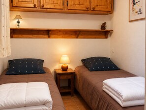 A bedroom with two beds, a nightstand with a lamp, wooden cabinets above, and a framed picture on the wall.