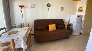 Een knus kamertje met een bruine bank, een eettafel met stoelen, een lamp, een klok en een koelkast.