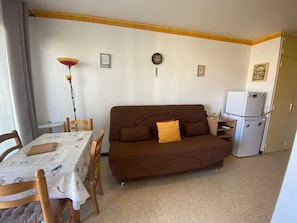 Une petite pièce agréable, avec un canapé marron, une table à manger entourée de chaises, une lampe, une horloge et un réfrigérateur.