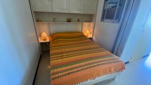 Een kleine, netjes ingerichte slaapkamer met een bed, twee bedlampen, een nachtkastje en een kastje boven het bed.