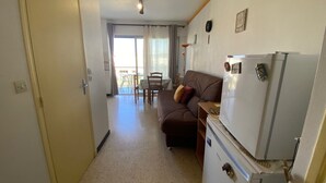 Een compacte woonruimte met een kitchenette, een bank, een eettafel met stoelen en een schuifdeur naar een buitenruimte.
