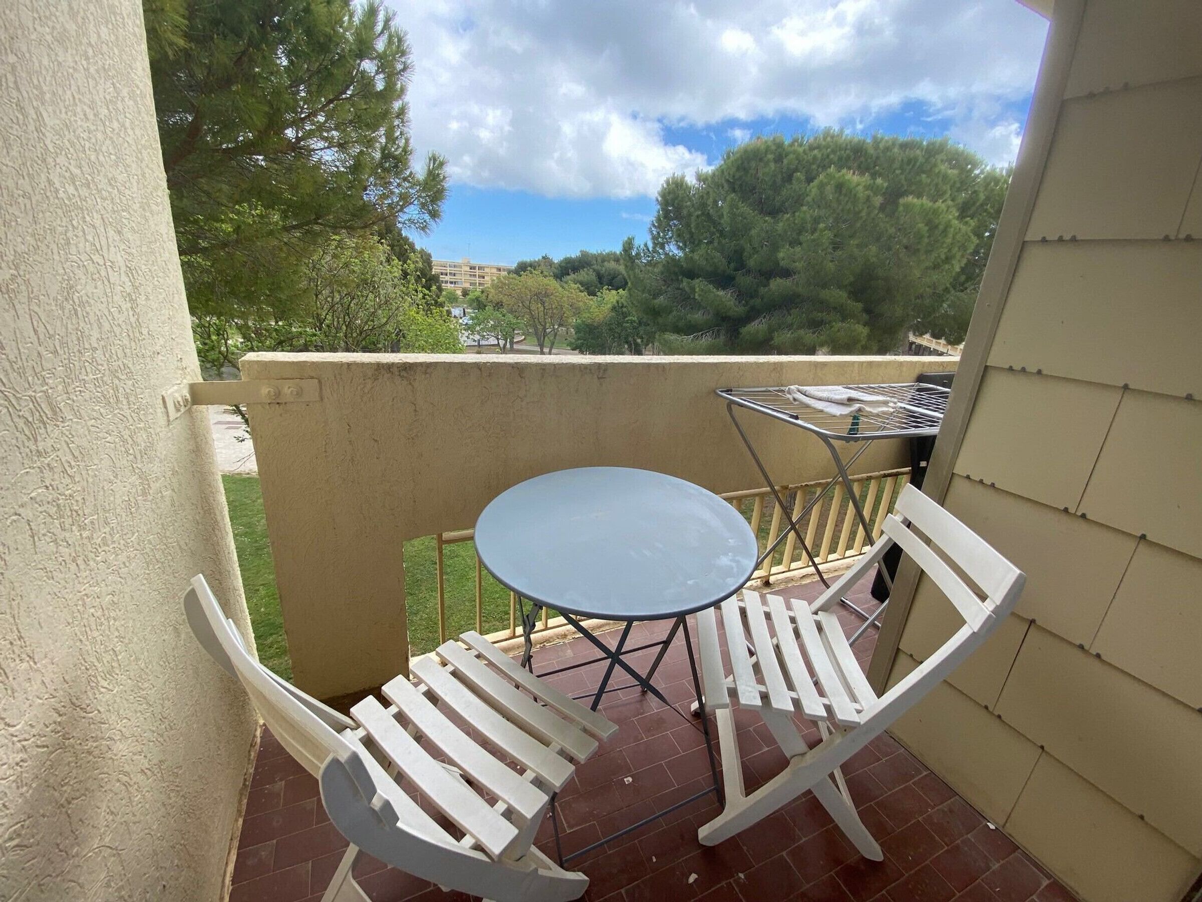 A balcony with a small table and two white chairs, a metal drying rack, and a view of trees and buildings in the background.