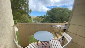 Een balkon met een kleine tafel en twee witte stoelen, een metalen rek en een uitzicht op bomen en gebouwen op de achtergrond.