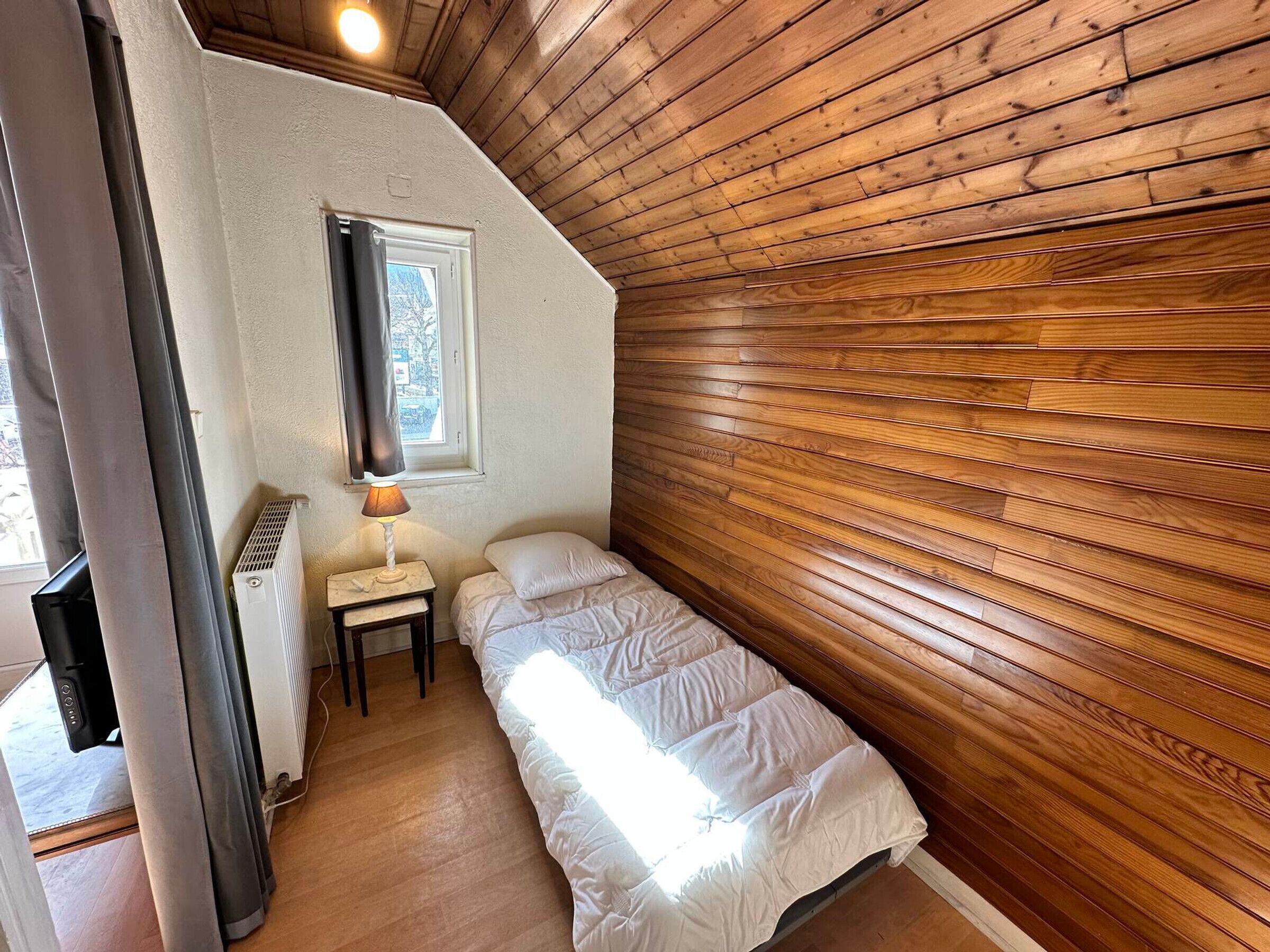 A small, cozy room with a sloped ceiling, a single bed, a bedside table with a lamp, and a window with curtains.