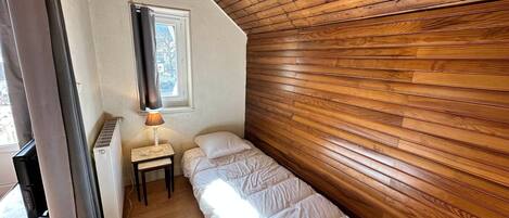 A small, cozy room with a sloped ceiling, a single bed, a bedside table with a lamp, and a window with curtains.