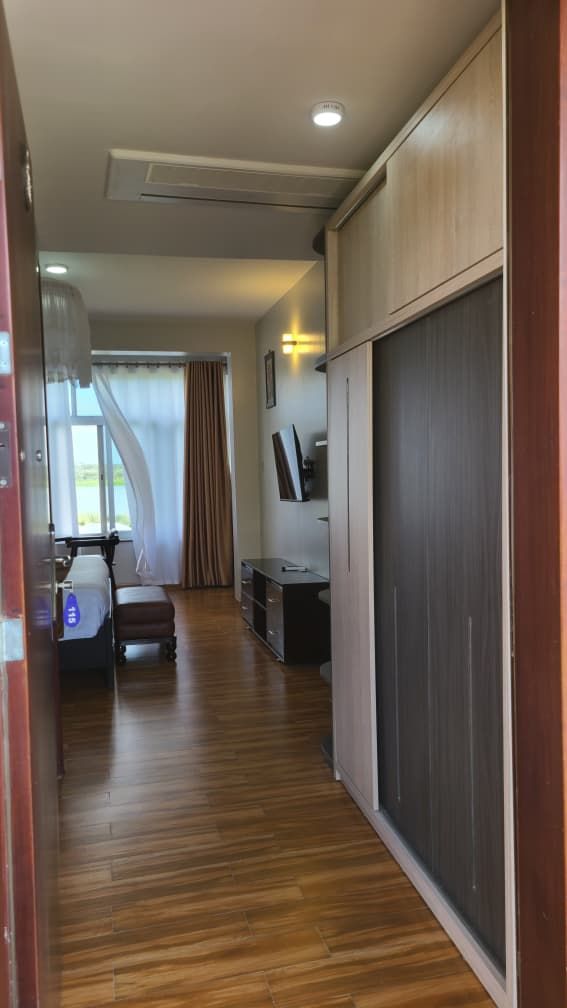 Un couloir doté d’un plancher en bois, d’une porte coulissante, d’un petit coin salon avec une chaise et une table, et d’une fenêtre avec des rideaux.