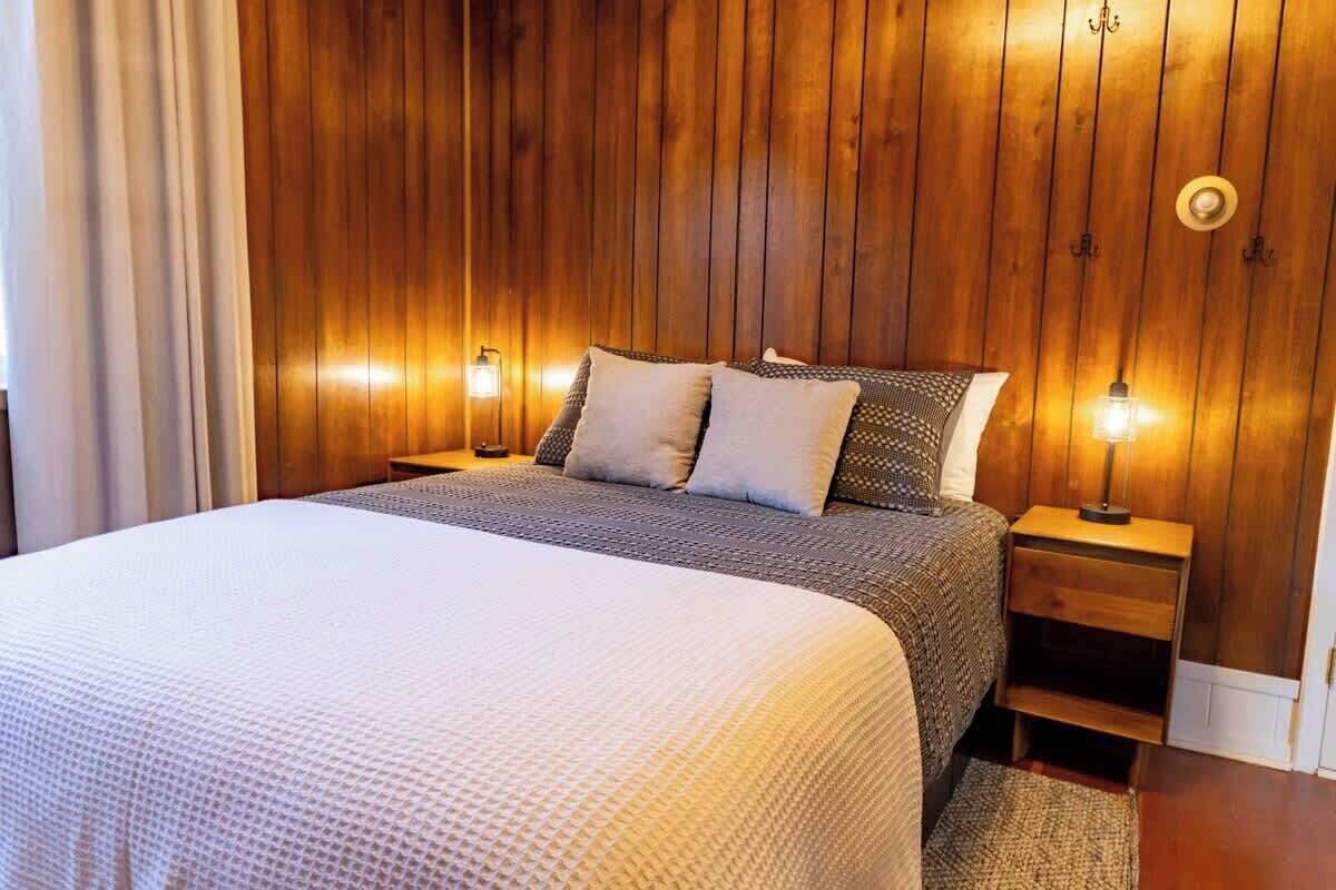 Un dormitorio con cabecera de madera panelada, una cama con colcha texturizada blanca, dos almohadas y lámparas de noche.