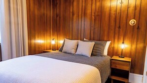 A bedroom with a wooden paneled headboard, a bed with a white textured bedspread, two pillows, and bedside lamps. - Hi-Bug Hideaway- In Town - Historic Home (Red Lodge)