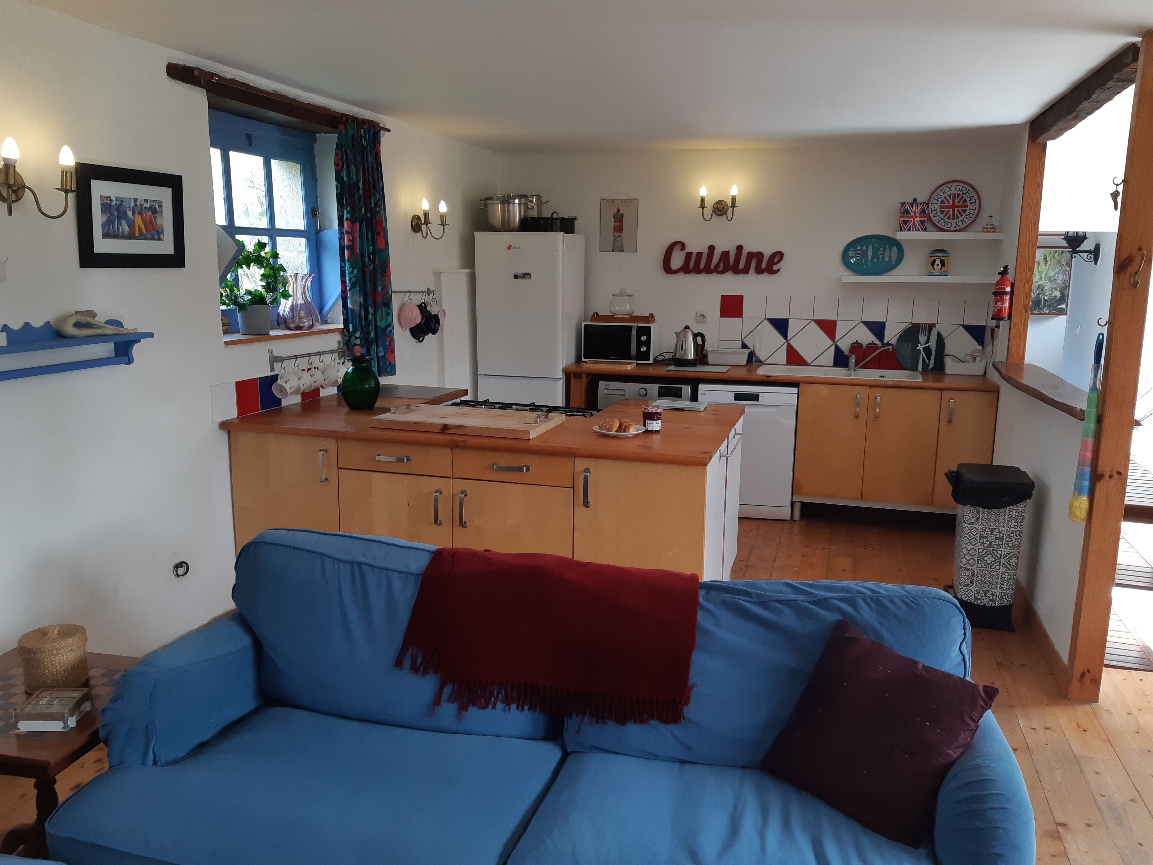 A cozy living room with a blue sofa and a red throw blanket, a kitchen with wooden cabinets and a white refrigerator, and a wall sign that reads "Cuisine".