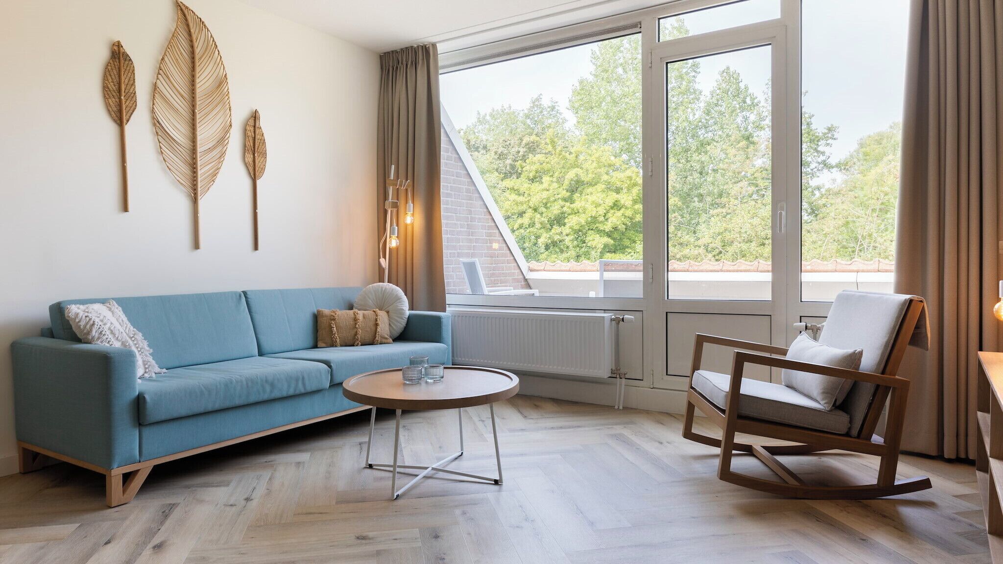 Ein modernes Wohnzimmer mit einem blauen Sofa, einem Holzschaukelstuhl, einem Couchtisch und großen Fenstern mit Blick auf Bäume.