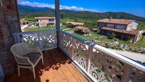 A wooden balcony with ornate white railings, a wicker chair, and a view of traditional buildings in a rural setting. - Anapea Village (Kvareli)