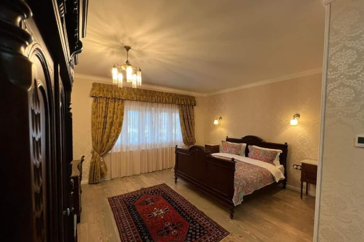 Un dormitorio con una cama de madera, un tapete con estampado rojo y negro, una araña y dos lámparas de pared.