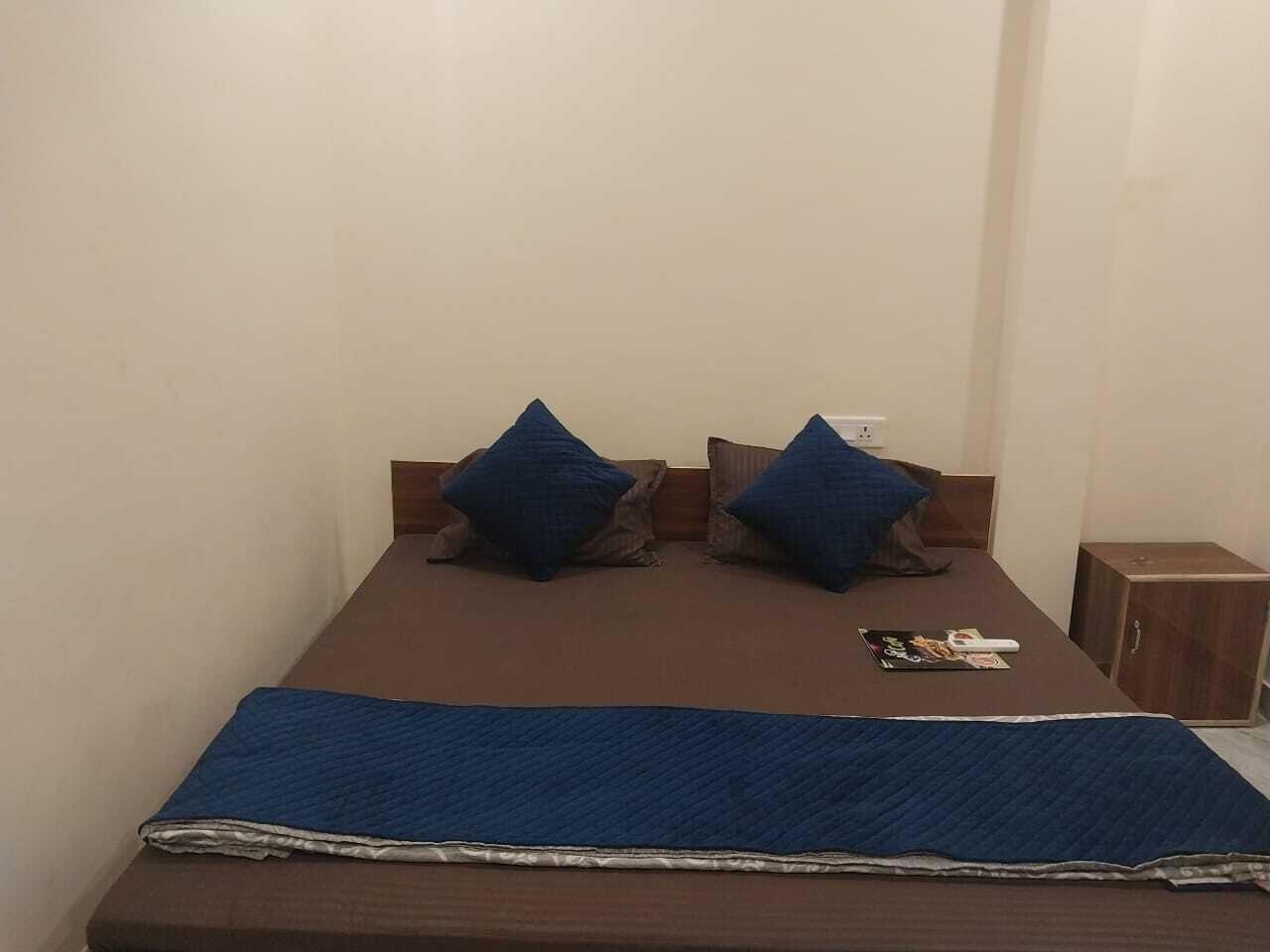 Una cama individual con una manta azul y dos almohadas, una mesita de noche de madera con un control remoto y un armario pequeño.